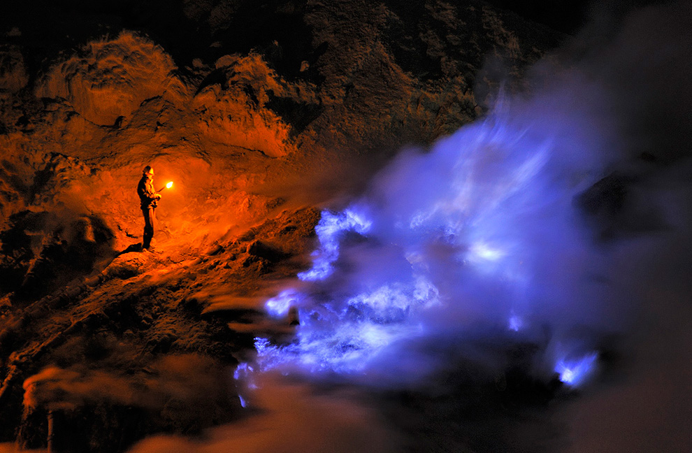 Шахтер стоит ночью в кратере Кава Иджен, держит факел и смотрит на поток жидкой серы, горящей синим пламенем.